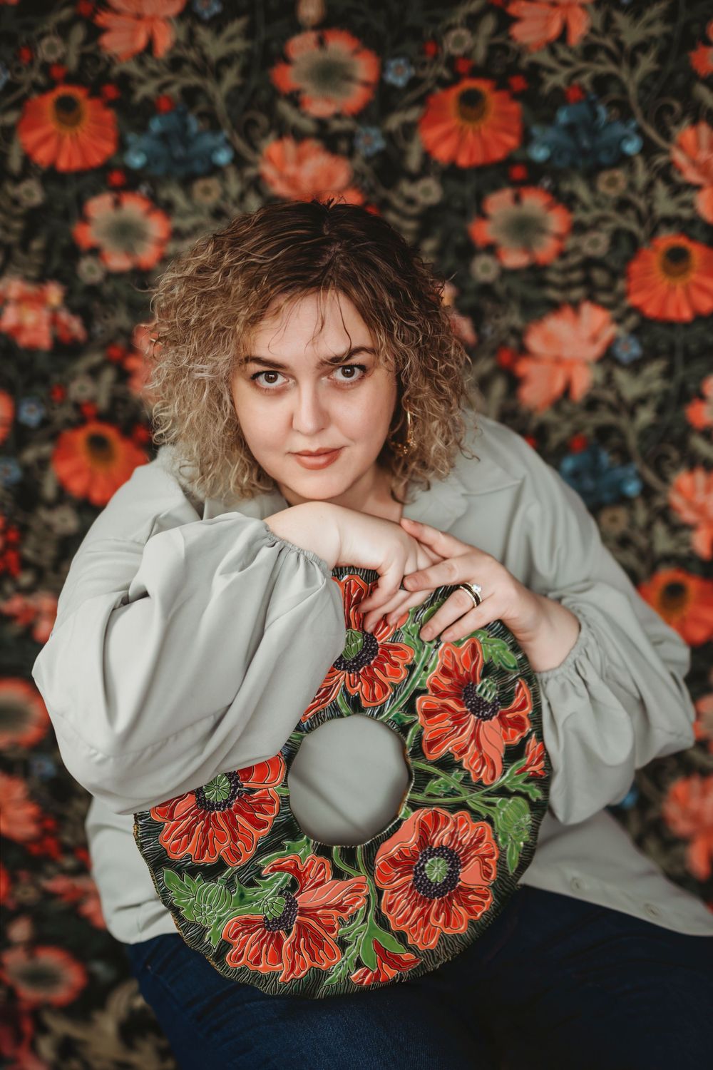 Curly-haired model poses with decorative floral wreath against orange poppy patterned wallpaper wearing light blouse.