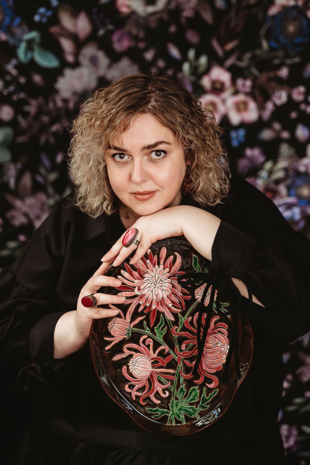 Artistic portrait in front of floral background with black dress holding embroidered decorative pillow with pink flower design.