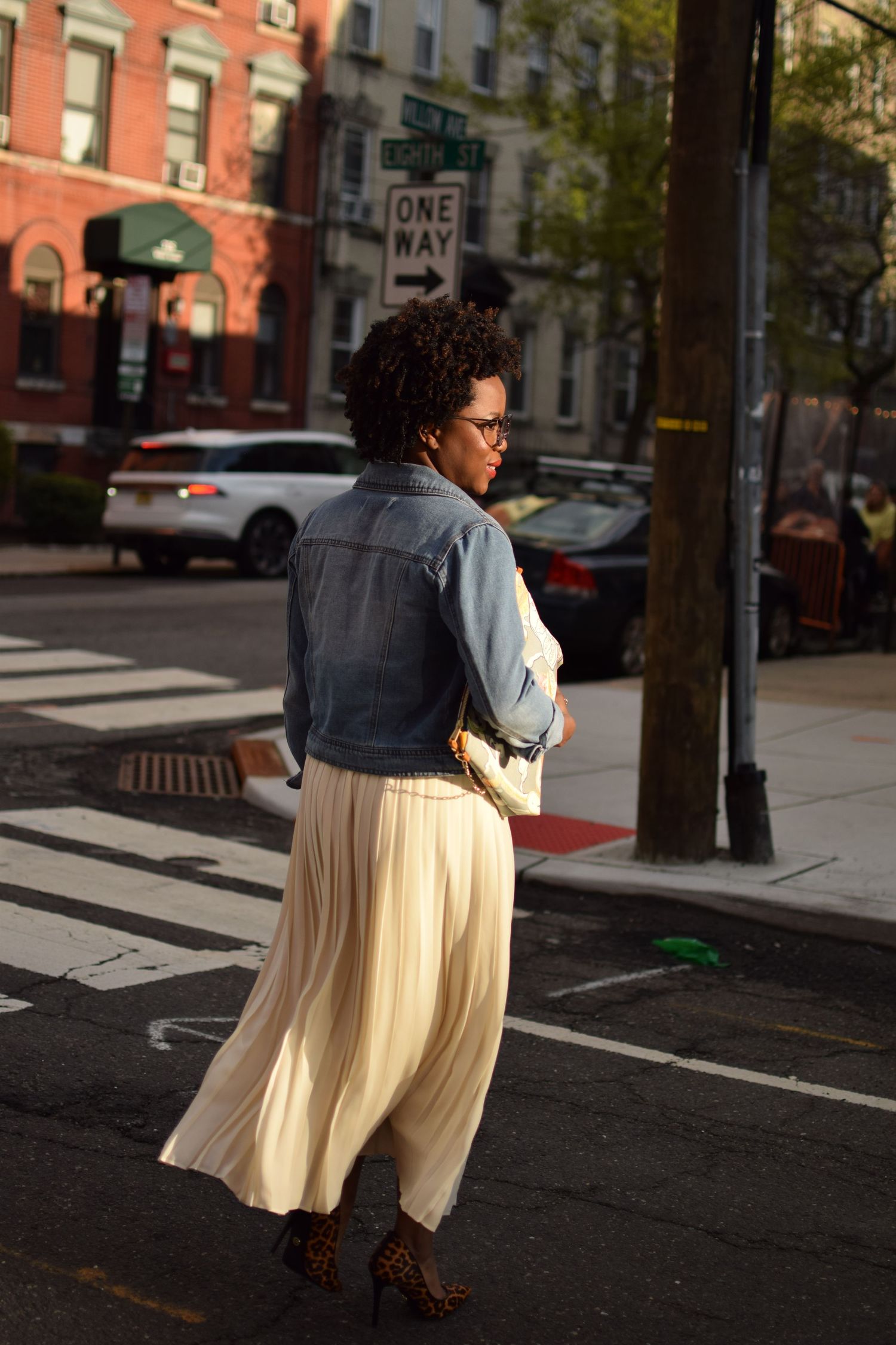A person in a denim jacket and pleated cream skirt stands on a sunny city street corner.