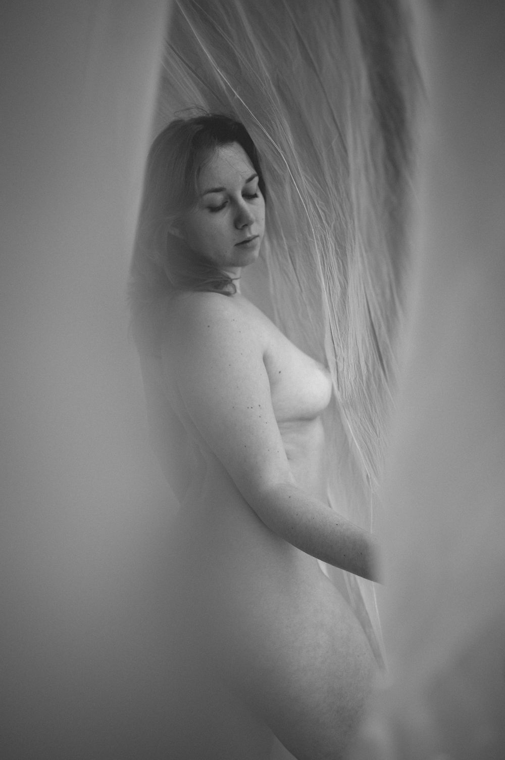 A soft black and white artistic nude figure study through sheer fabric creating ethereal silhouette effect.