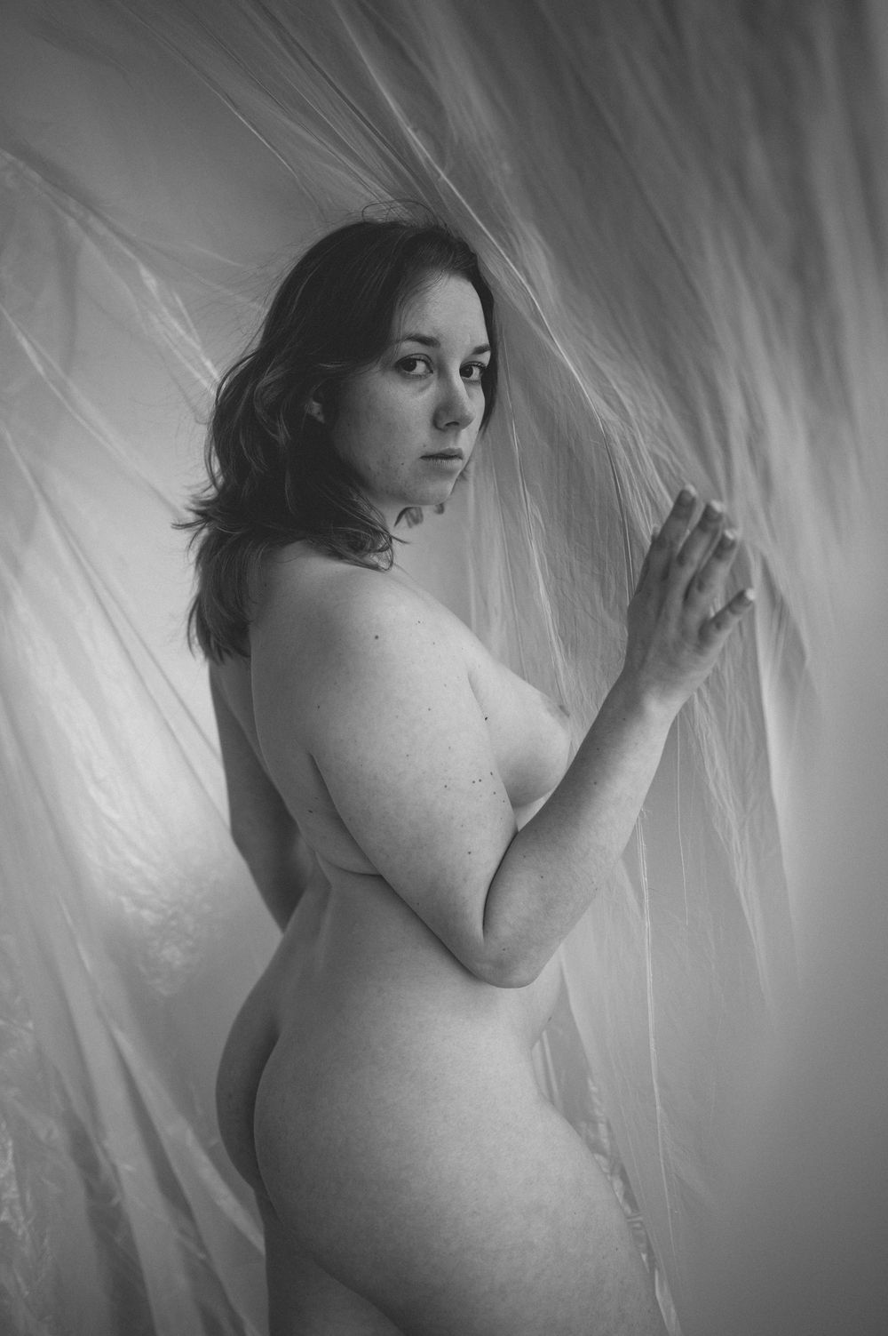Black and white artistic nude photograph featuring feminine silhouette against flowing fabric backdrop in soft lighting.