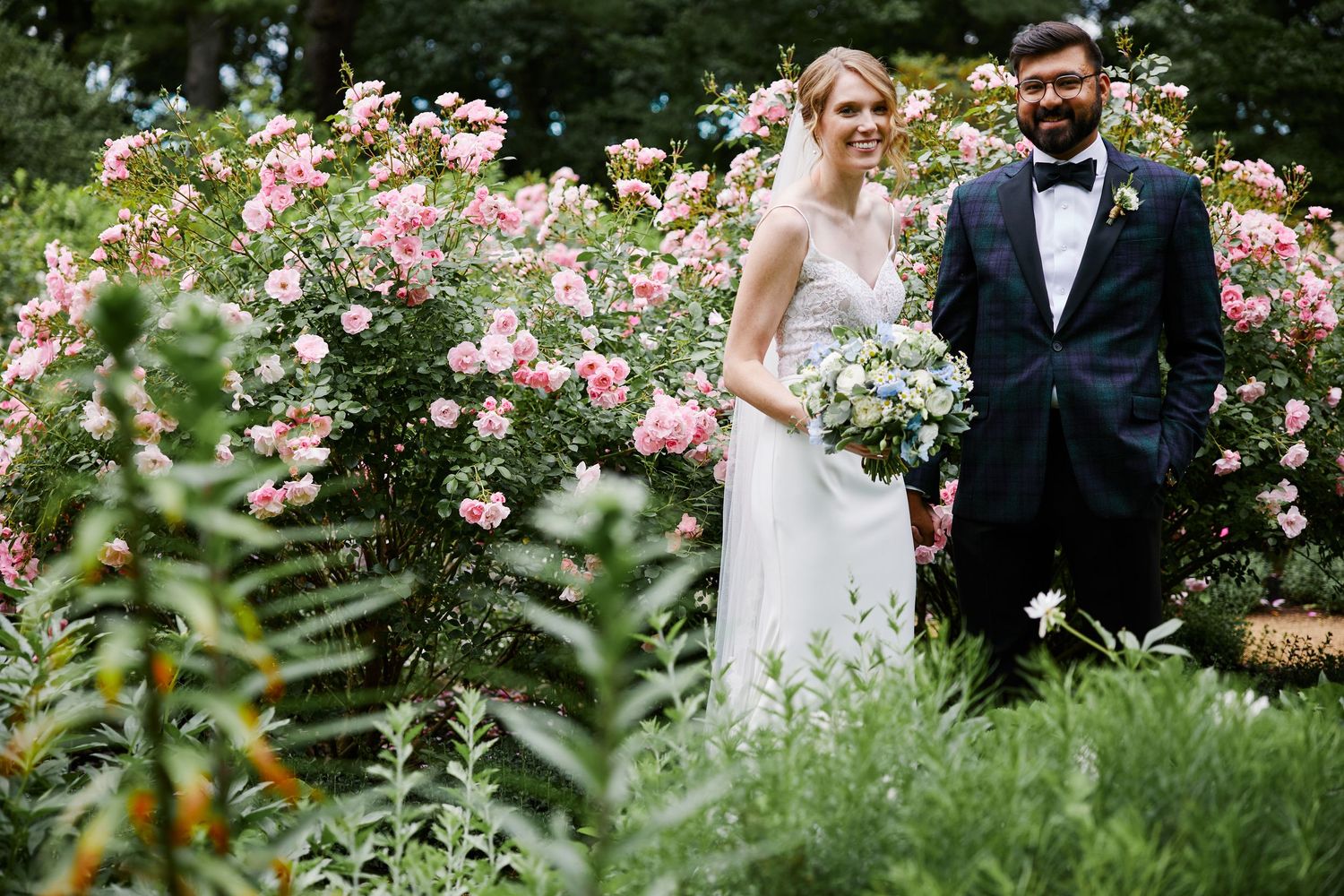 A couple poses in a romantic garden setting surrounded by blooming pink roses and lush greenery on their wedding day.