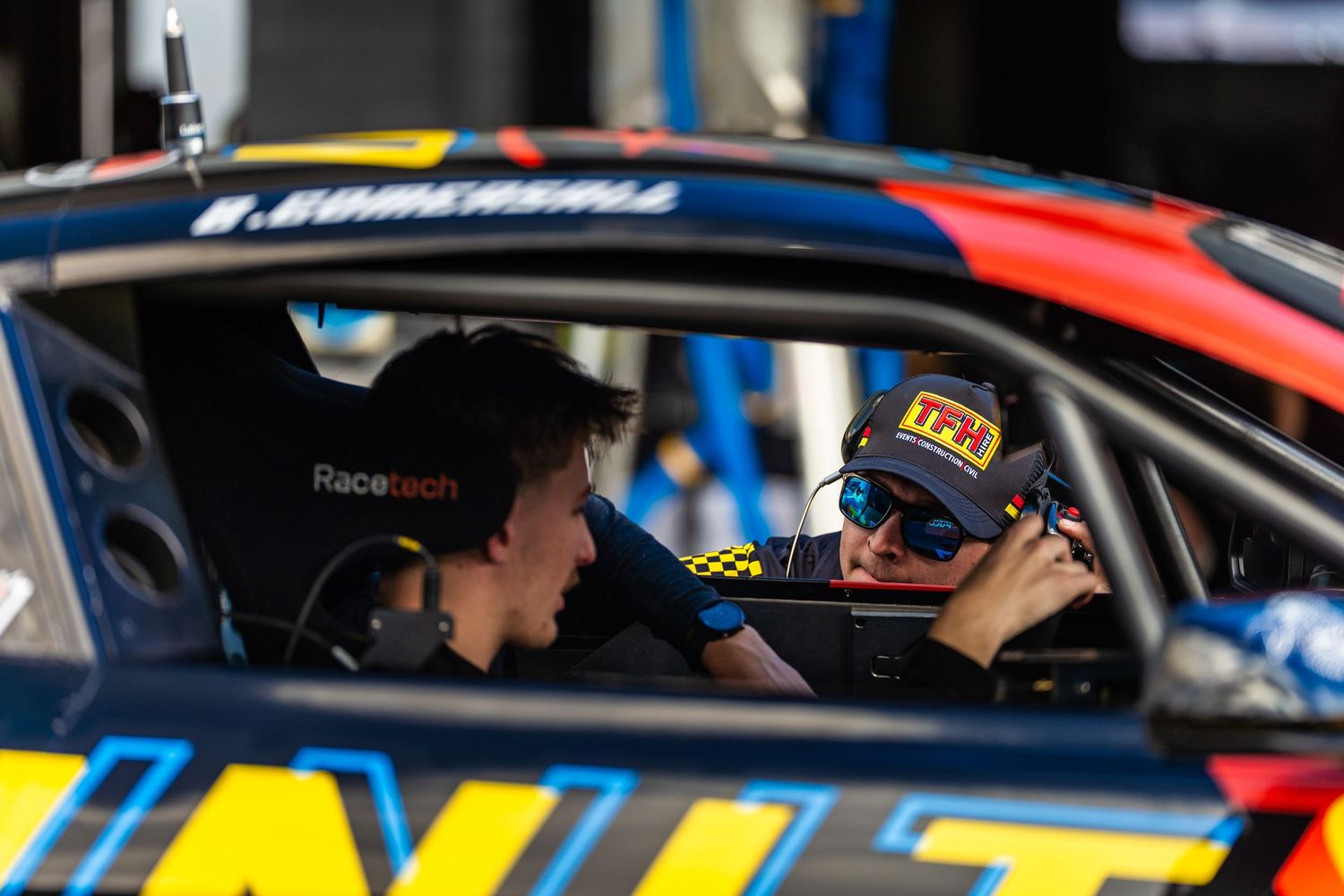 Two people in conversation sit inside a race car visible through the driver's side window.