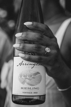 A hand with white nails and diamond ring holds a bottle of Arrington Riesling wine.