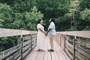 A couple holds hands while standing on a wooden bridge surrounded by lush green trees.