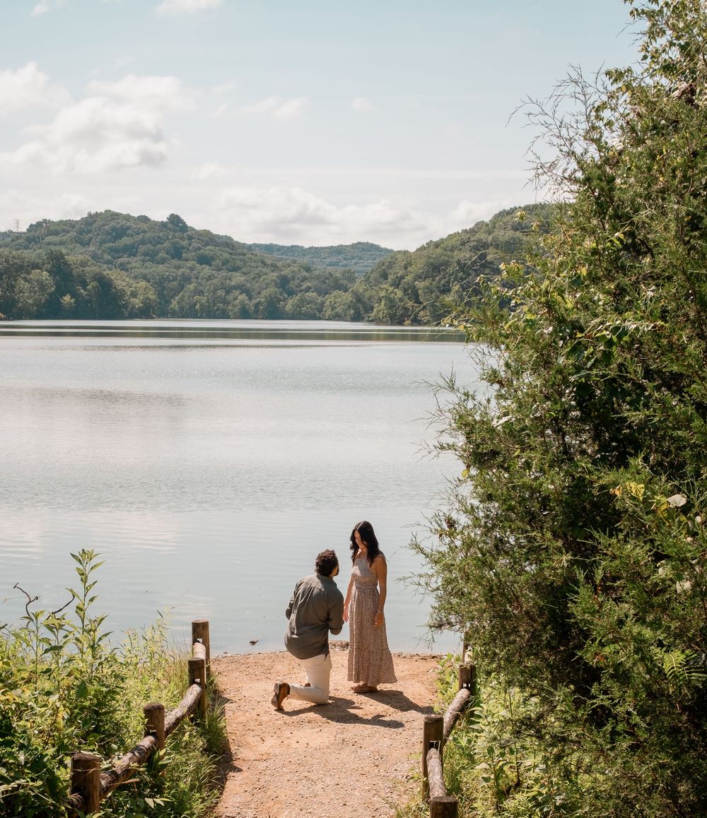 Two people stand on a wooden dock overlooking a serene lake surrounded by lush green hills on a tranquil afternoon.