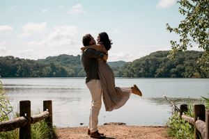 A couple shares a romantic embrace by a scenic lake surrounded by rolling hills and wooden fence posts in warm sunlight.