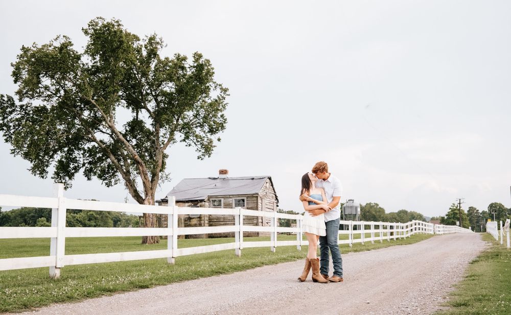 A couple embraces near a white fence on a country path with a large tree and farmhouse in the background.