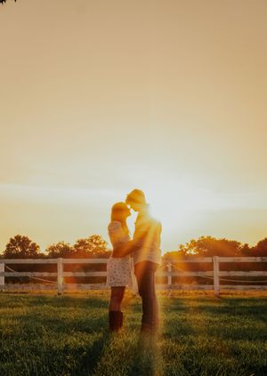 Couple embraces in a grassy field at sunset with white fence in background creating silhouette effect.