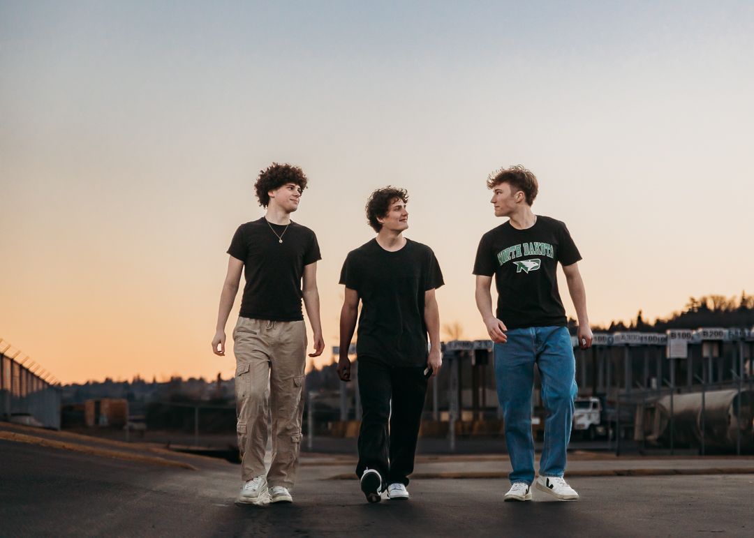 Three young people in casual attire walking together on a road at sunset with buildings in the background.