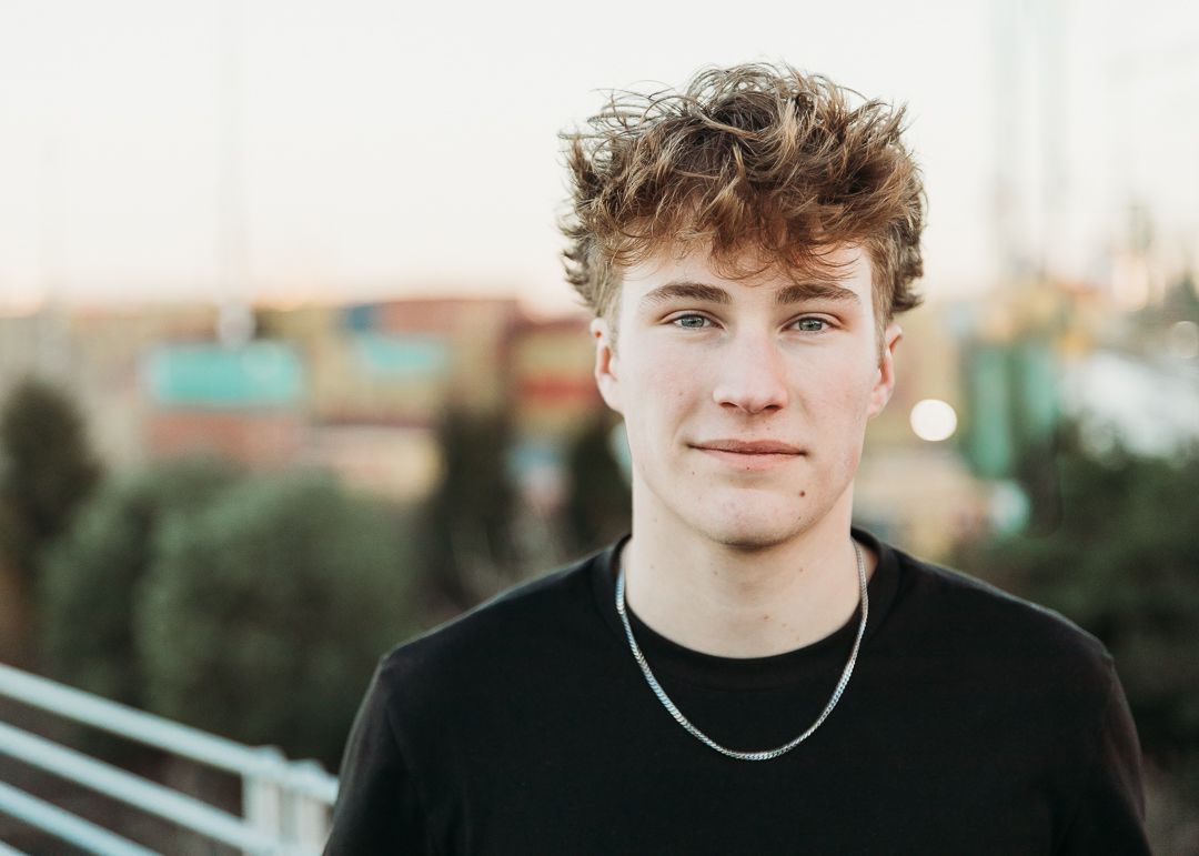 Person with messy brown hair wearing a black t-shirt and silver necklace poses outdoors at sunset.