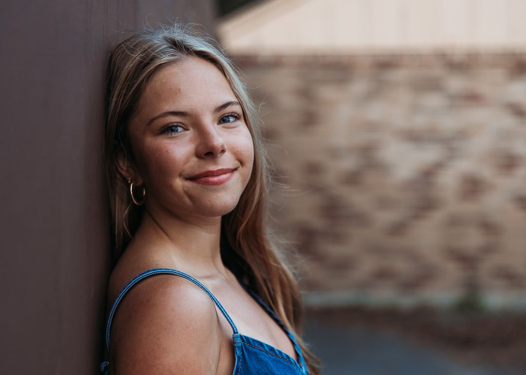 A young woman in a blue tank top smiles warmly while posing against a textured stone wall.