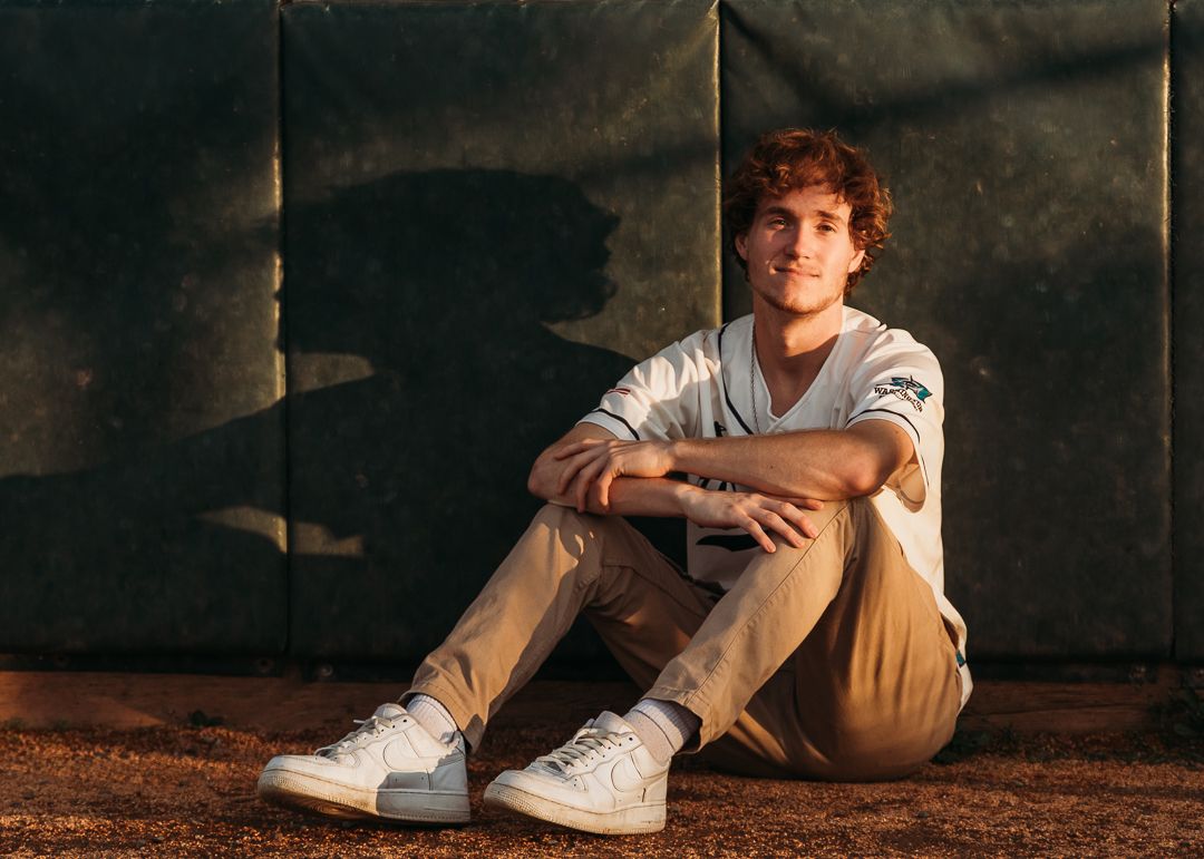 Someone wearing khaki pants and white sneakers sits casually against a dark textured wall in warm lighting.