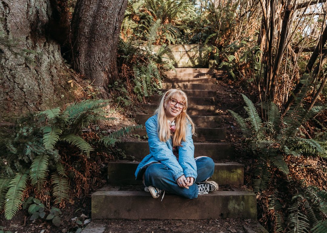 A person in blue sits on stone steps surrounded by lush ferns and trees in a forest setting.