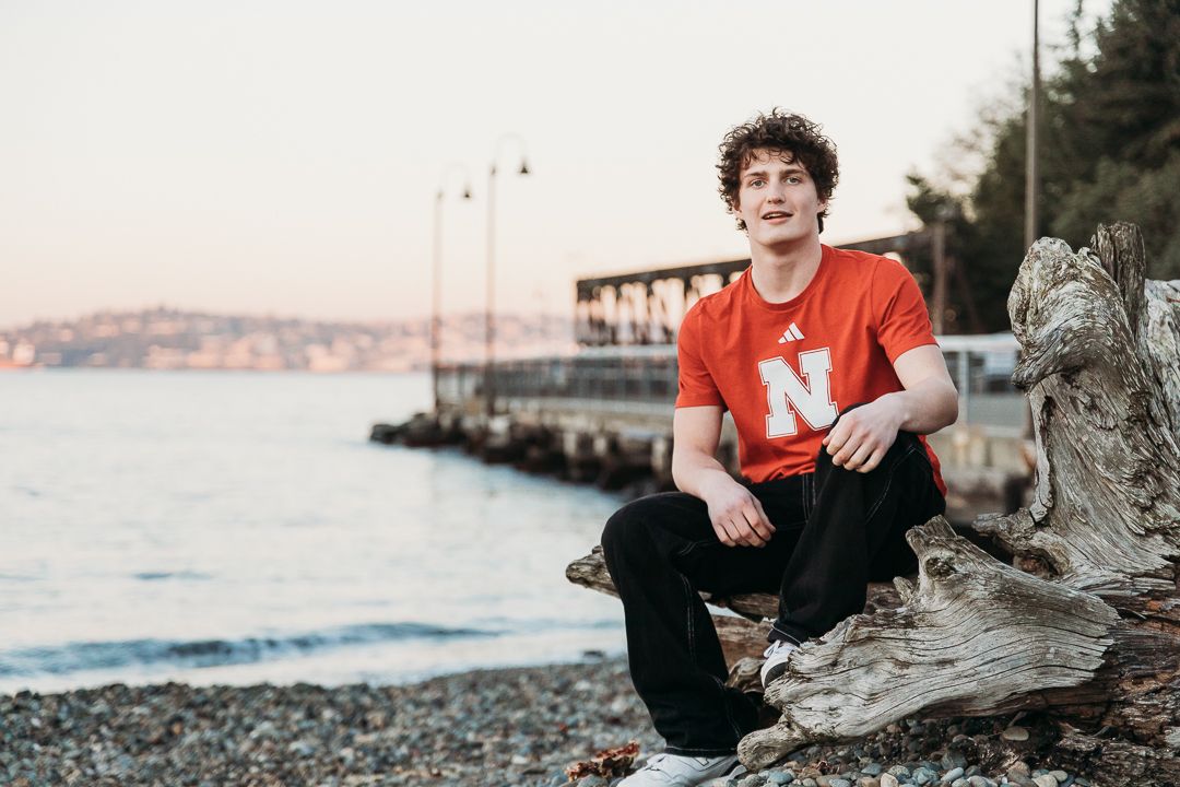 A person in a red shirt sits casually on rocky shore with a waterfront dock and distant coastline in the background.