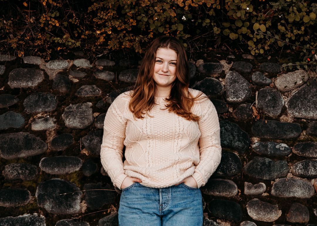 A person in a cream sweater poses against a stone wall during an outdoor photoshoot.