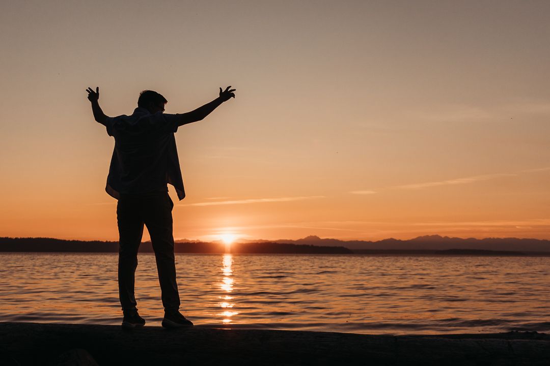 Silhouette of person with raised arms stands on shoreline during golden sunset over calm water and mountain landscape.