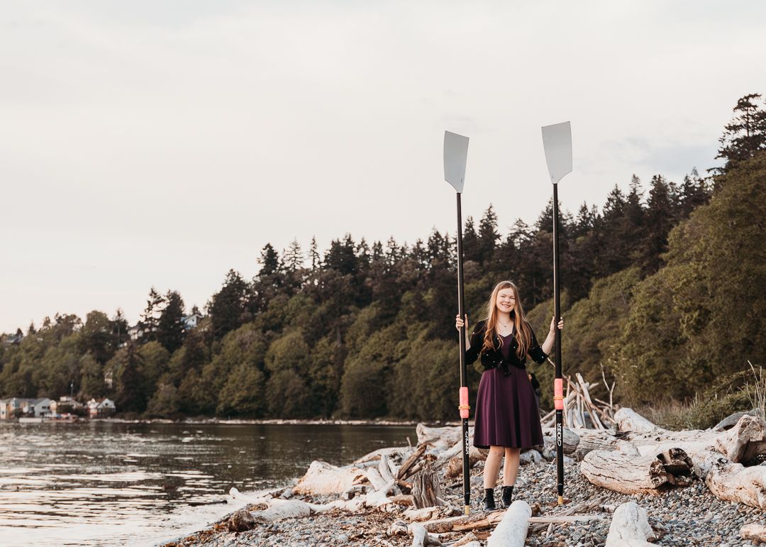 A person in a black dress stands on a rocky beach shoreline holding rowing oars against a forested backdrop.
