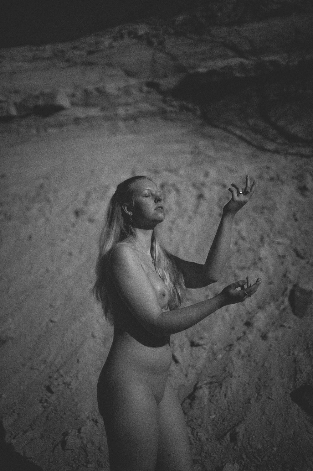 Artistic black and white nude photograph of figure posing expressively against rocky desert landscape backdrop.