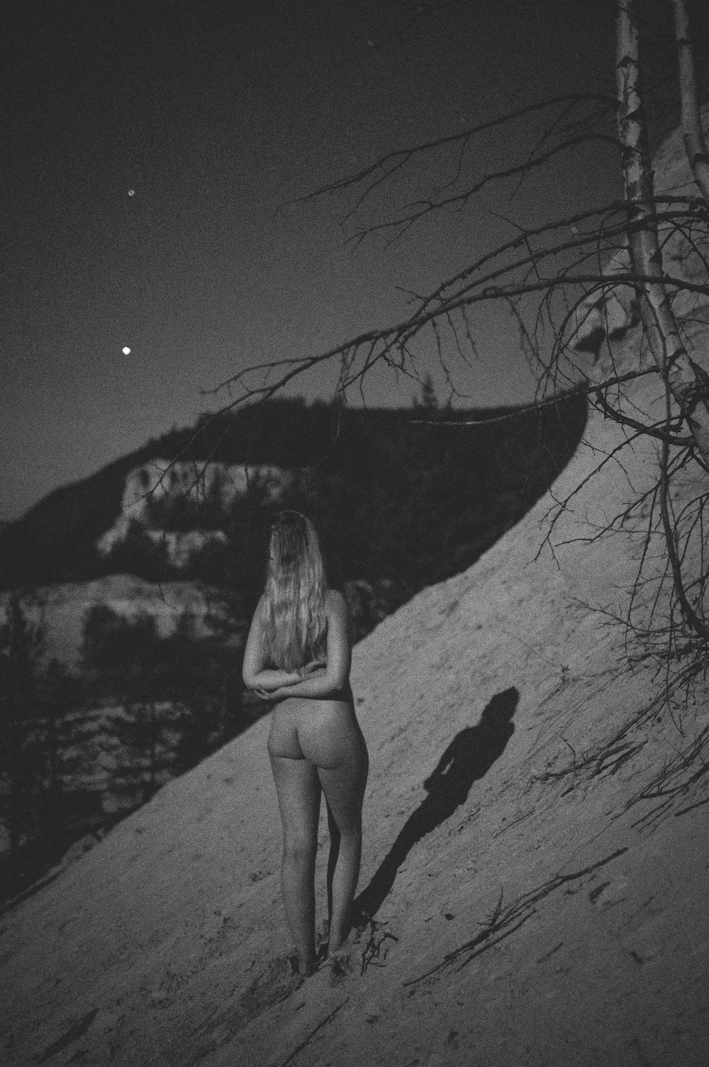 A silhouette against a mountainous backdrop stands on a rocky slope at dusk in black and white.