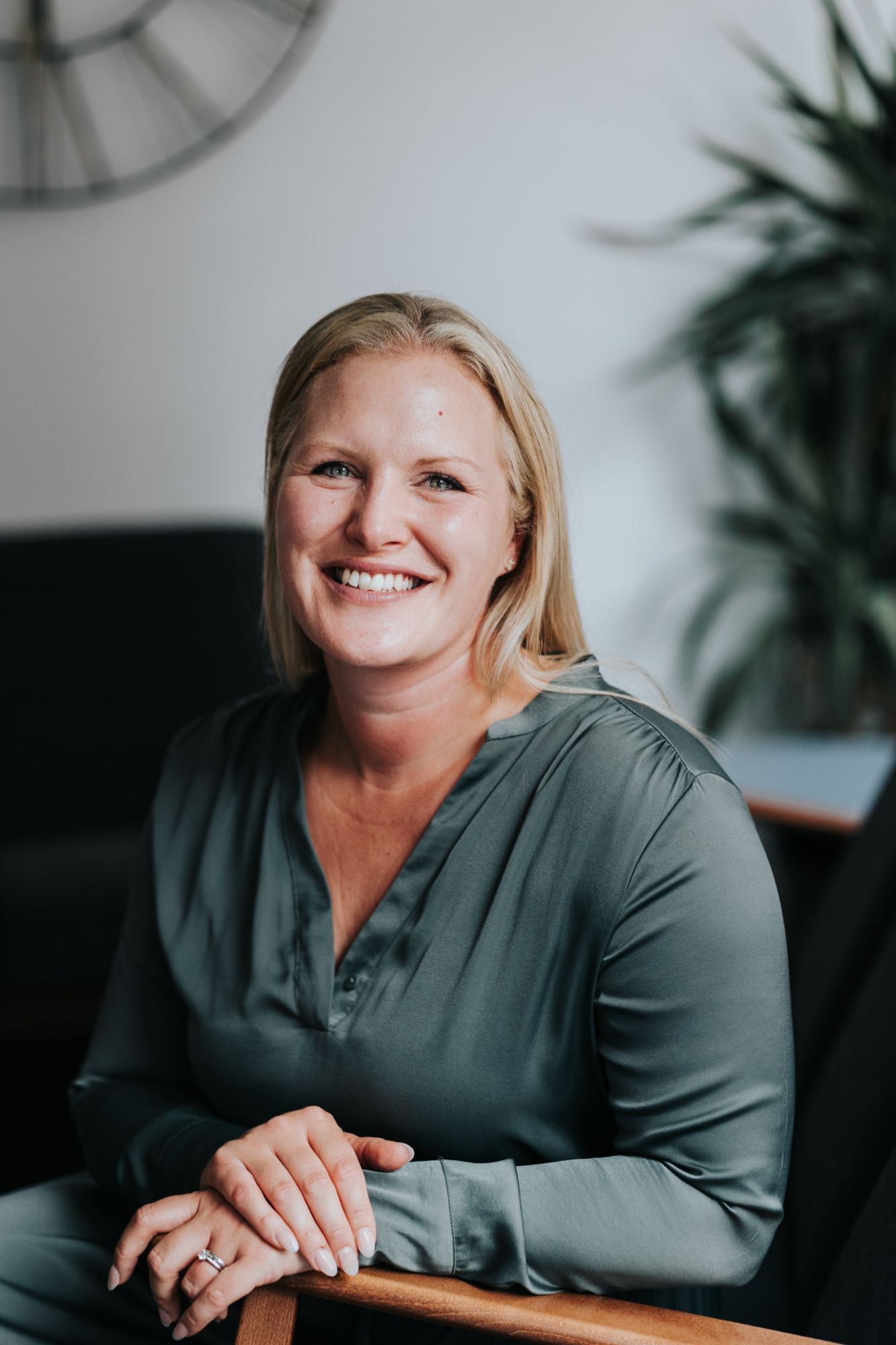 Professional headshots of person in gray blouse laughing against white wall with clock and plant decor.
