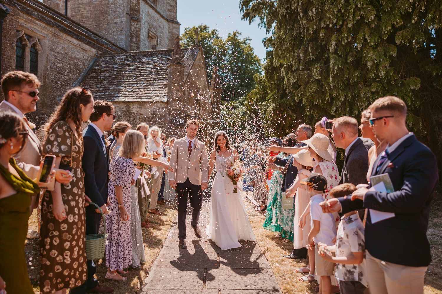 Wedding guests celebrate outside a historic stone church by throwing confetti on a sunny summer day.