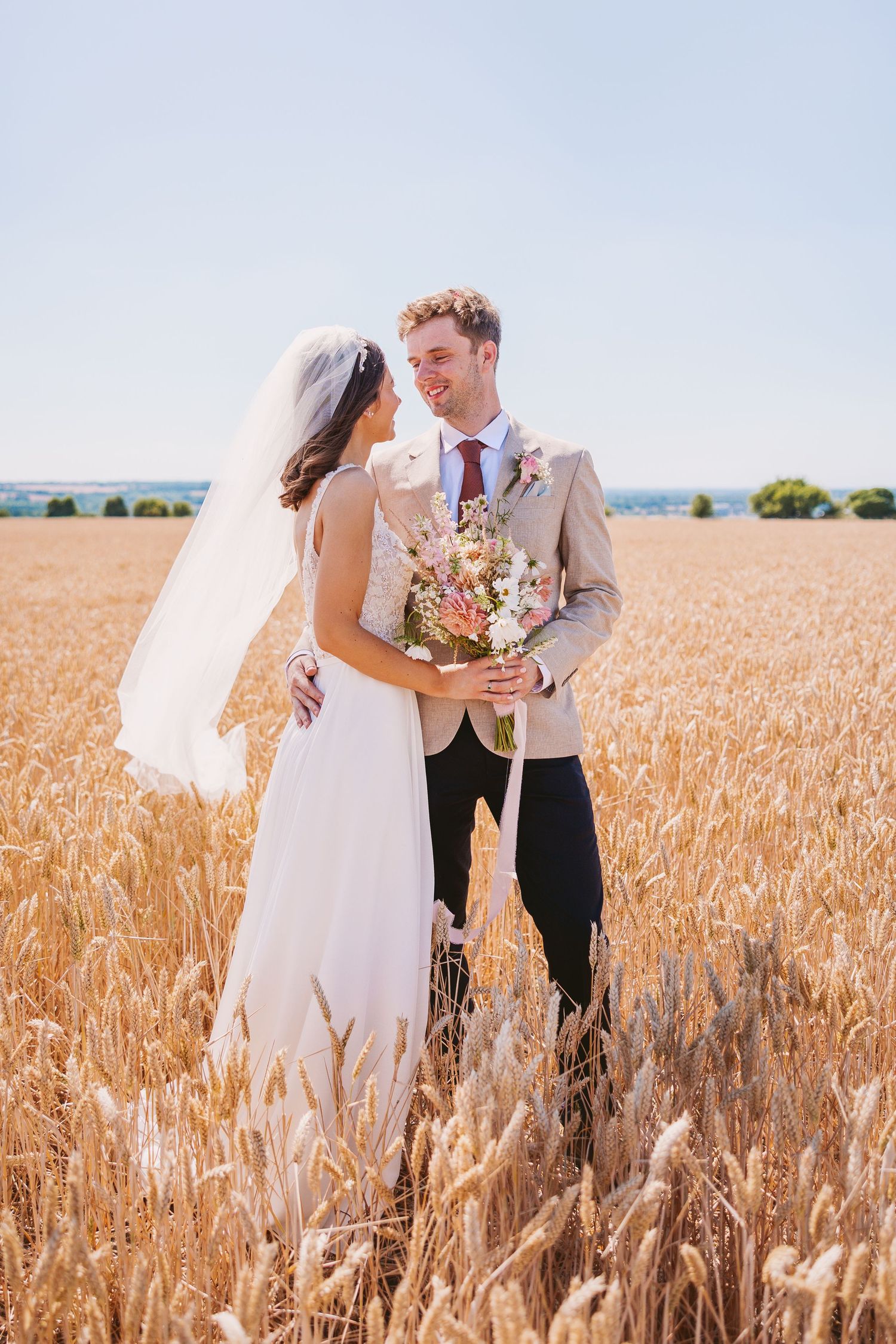 A romantic wedding couple stands together in a golden wheat field holding a bouquet on a sunny summer day.