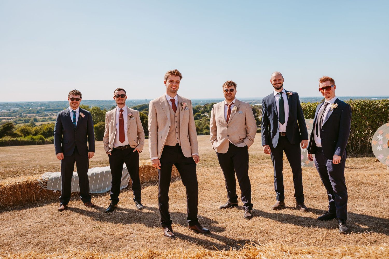 Groomsmen in formal attire stand together on a hilltop overlooking a scenic landscape on a sunny wedding day.