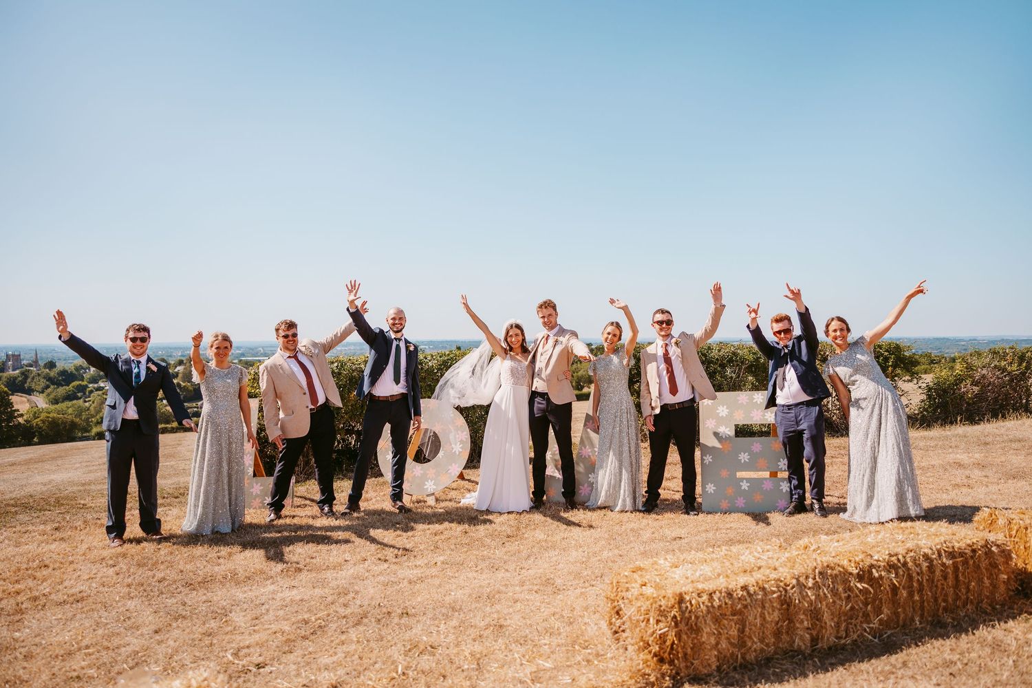 Wedding party members celebrate with raised arms against a scenic blue sky backdrop during an outdoor celebration.
