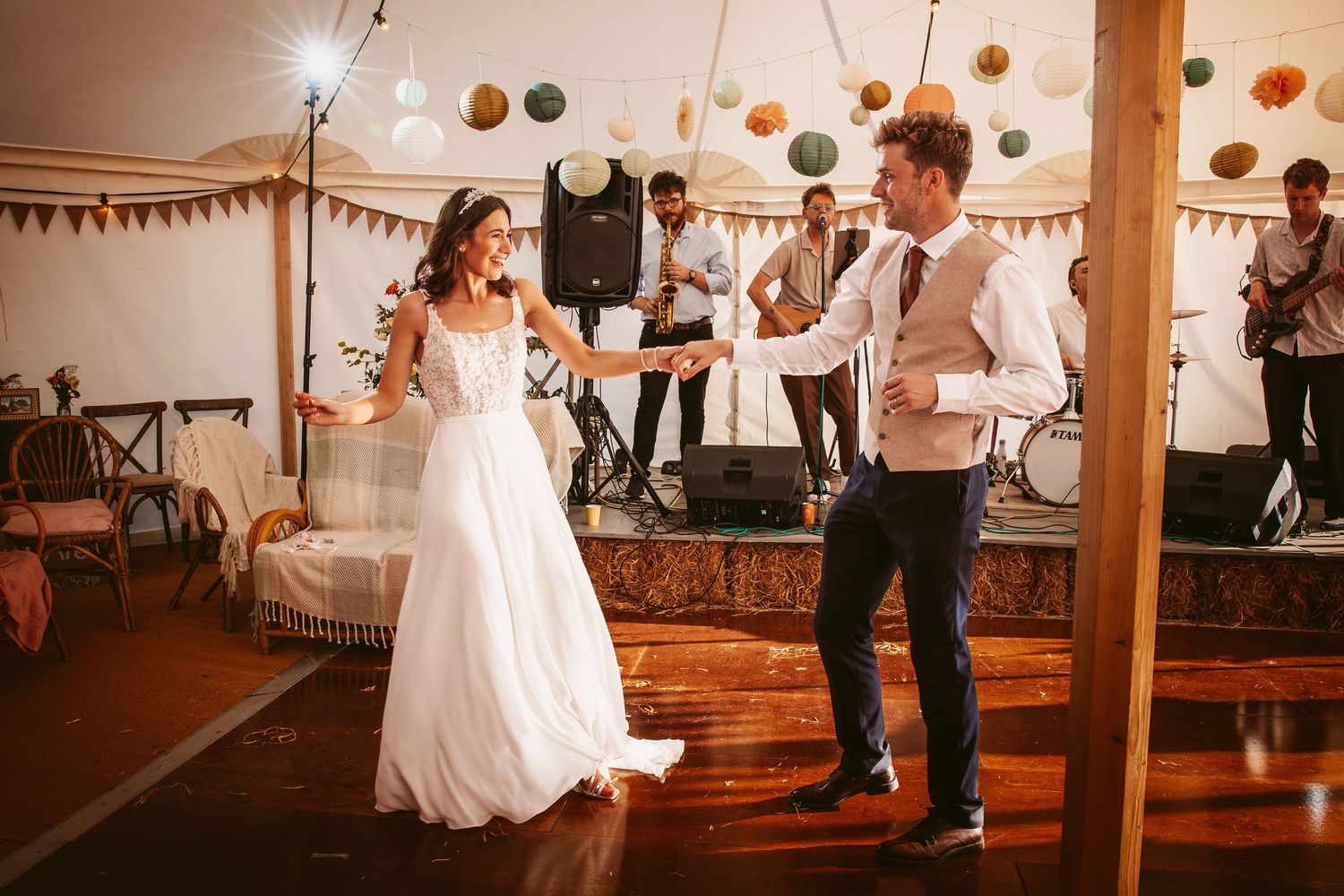 Wedding party dances and celebrates under hanging paper lanterns and string lights in a rustic indoor venue.