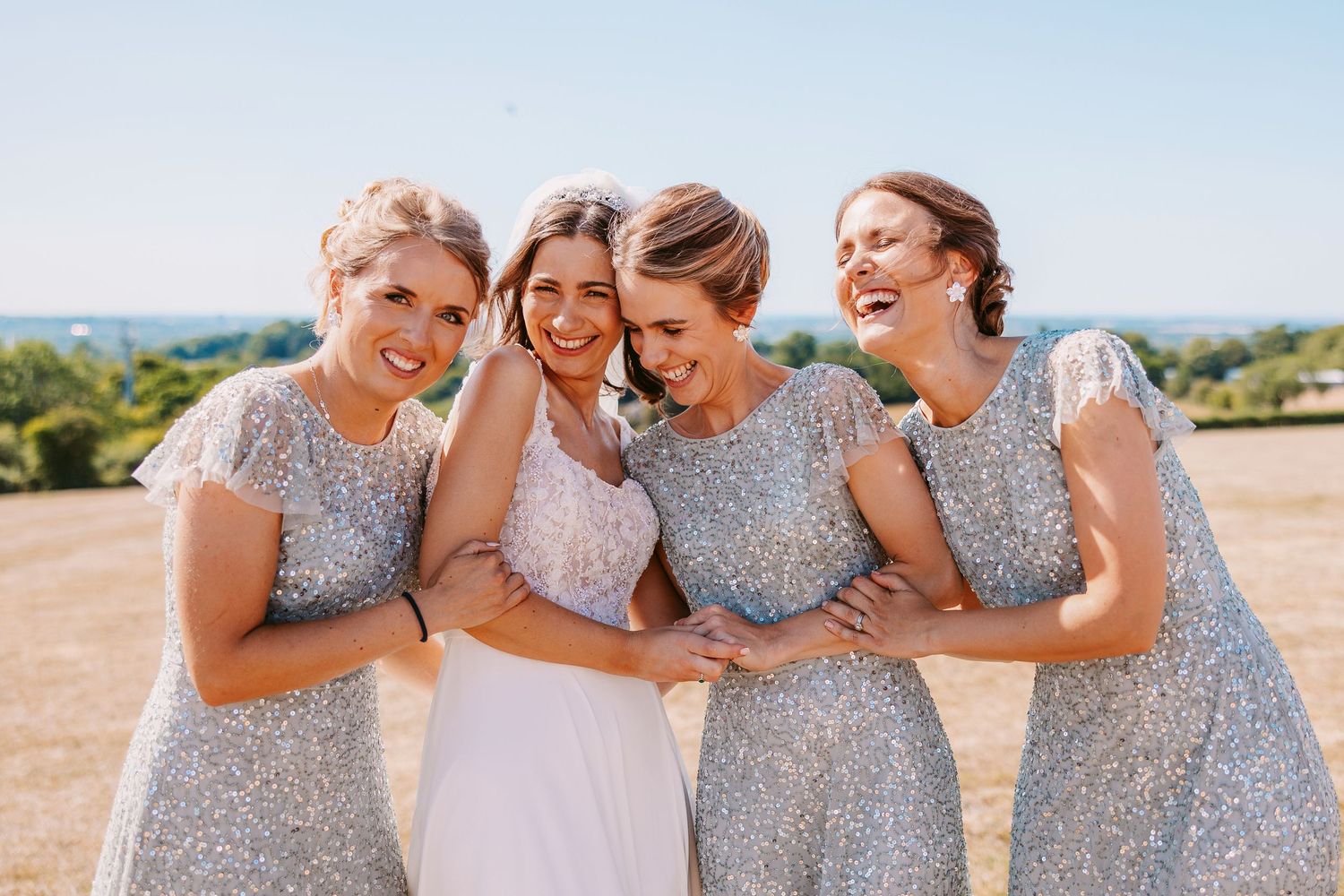Bridesmaids wearing sparkly silver dresses laugh together during outdoor wedding photo session.