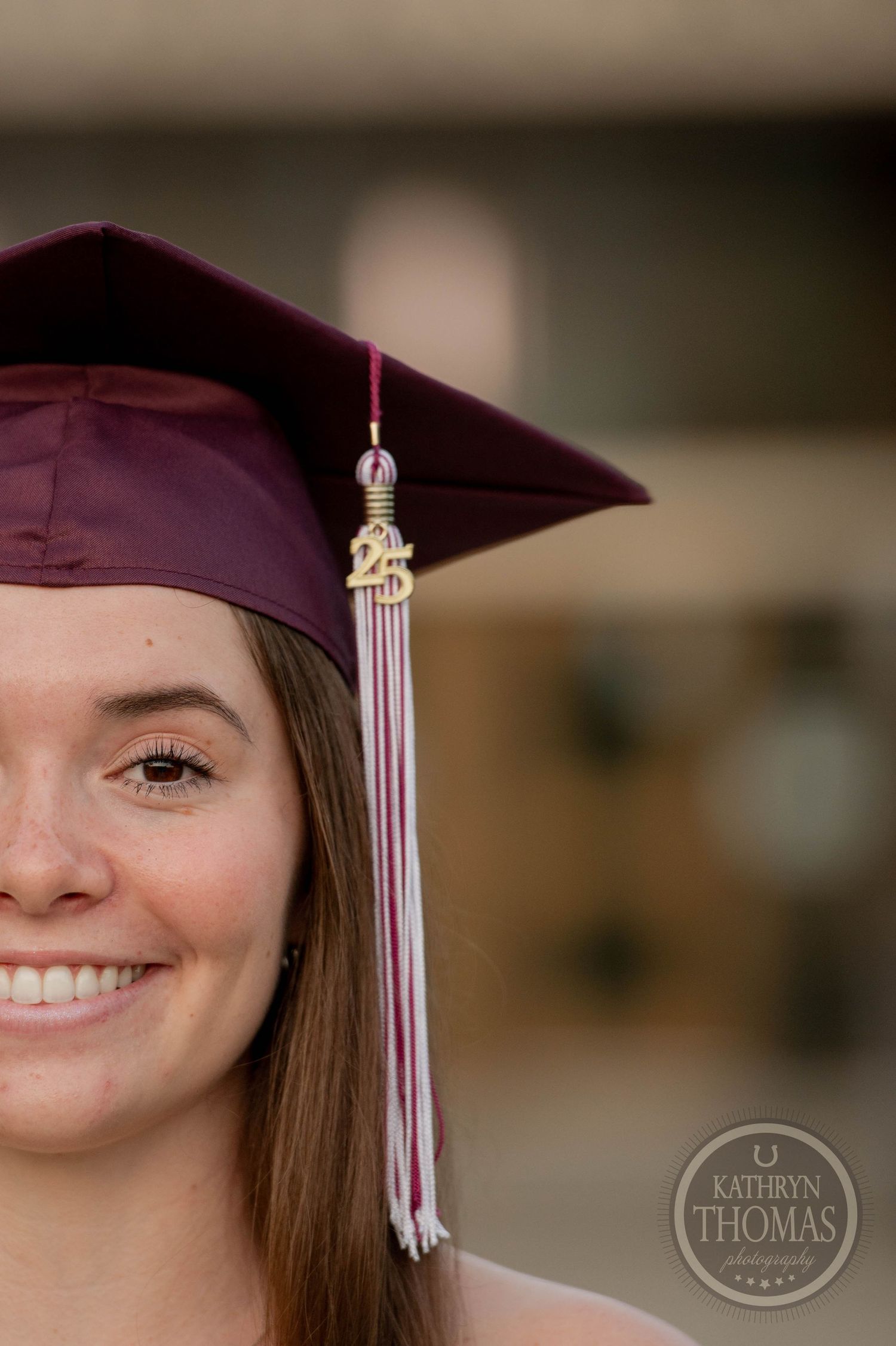 Graduate wearing burgundy cap with silver decorative tassel smiles in portrait photo by Kathryn Thomas Photography.
