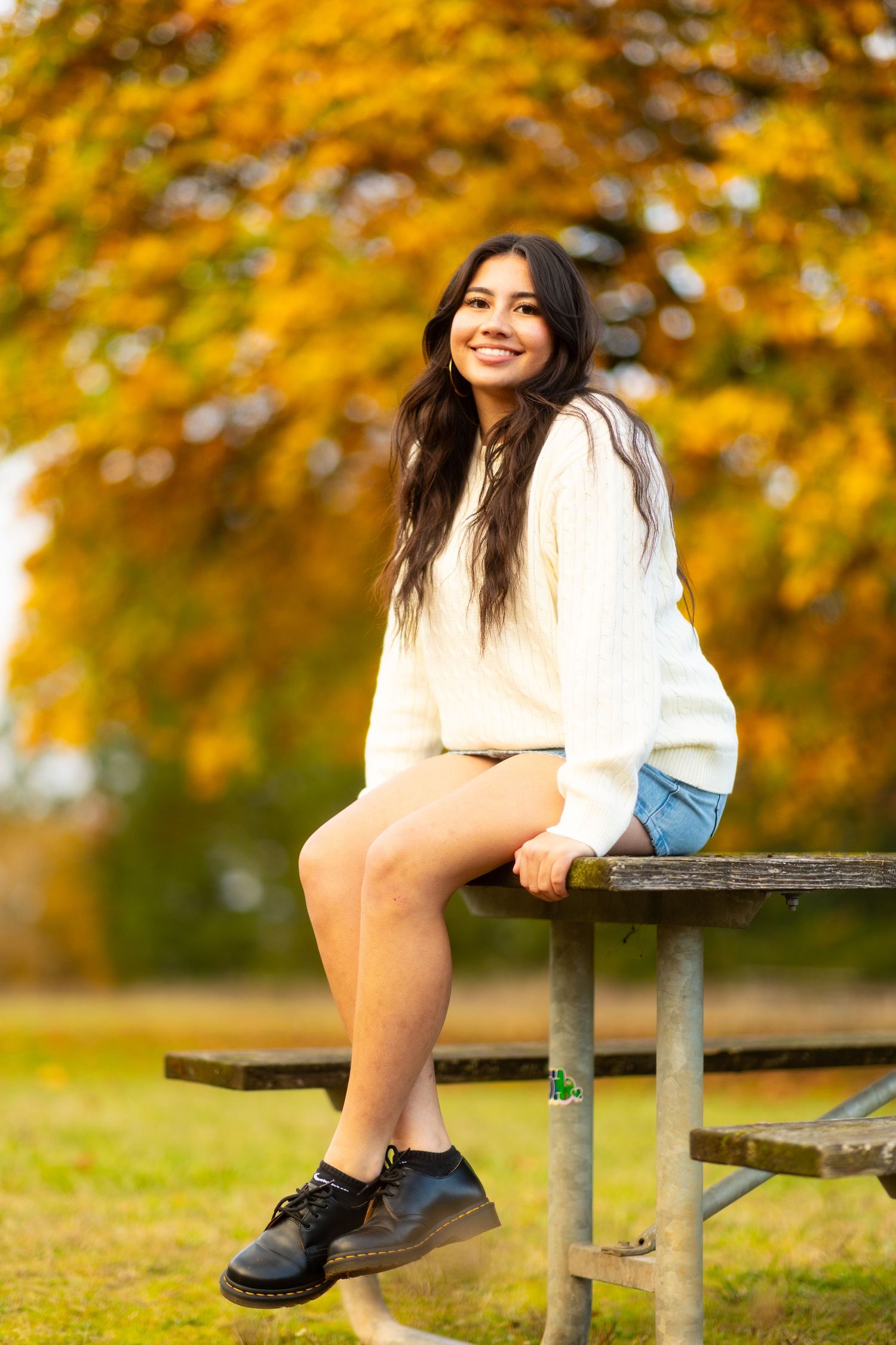 Someone in a white sweater and shorts sits on a picnic bench surrounded by golden autumn foliage in a park.