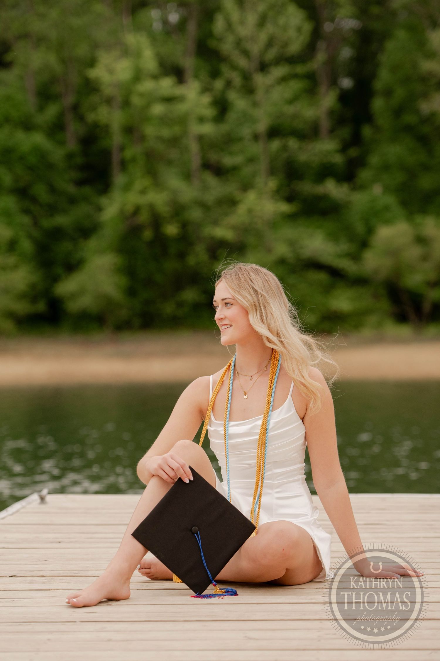 A graduate in a white swimsuit sits on a wooden dock by a lake holding their diploma, surrounded by green trees.