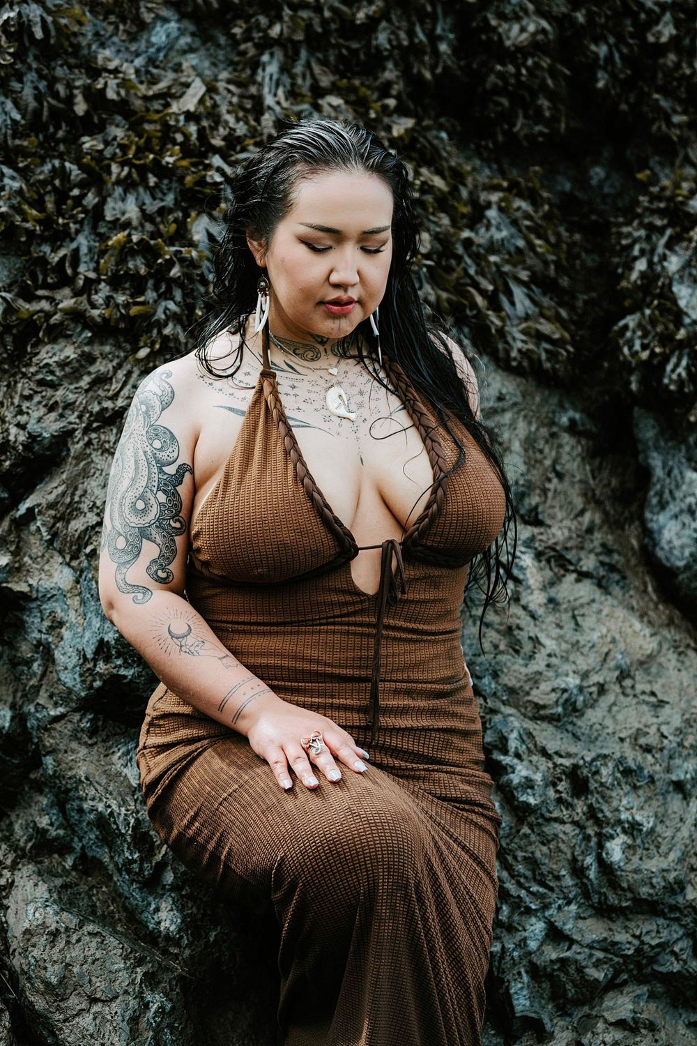 A woman in a brown dress poses against a rocky background while showing her tattoos and curves.