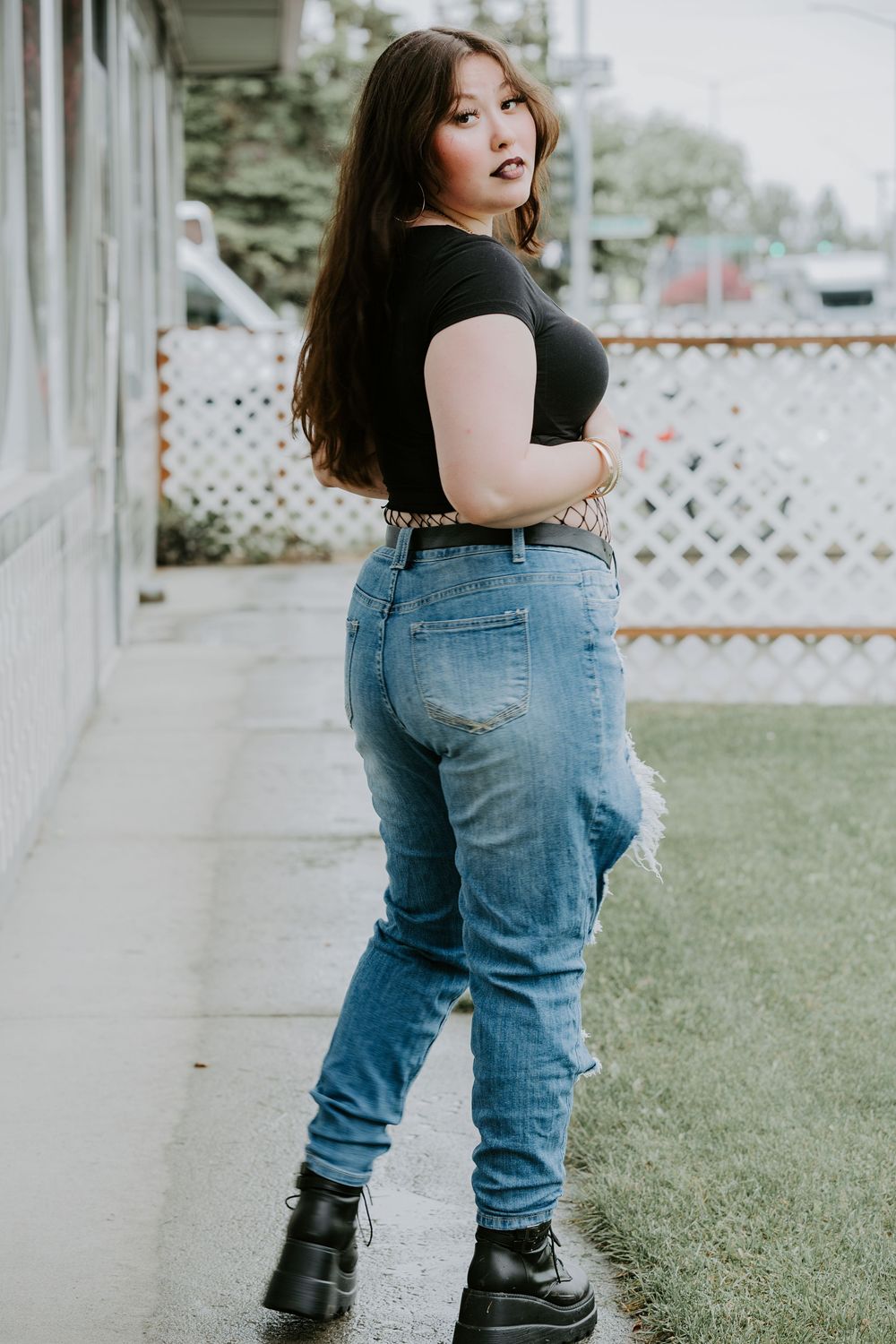 A woman in a black shirt and blue jeans poses against a white lattice fence backdrop.