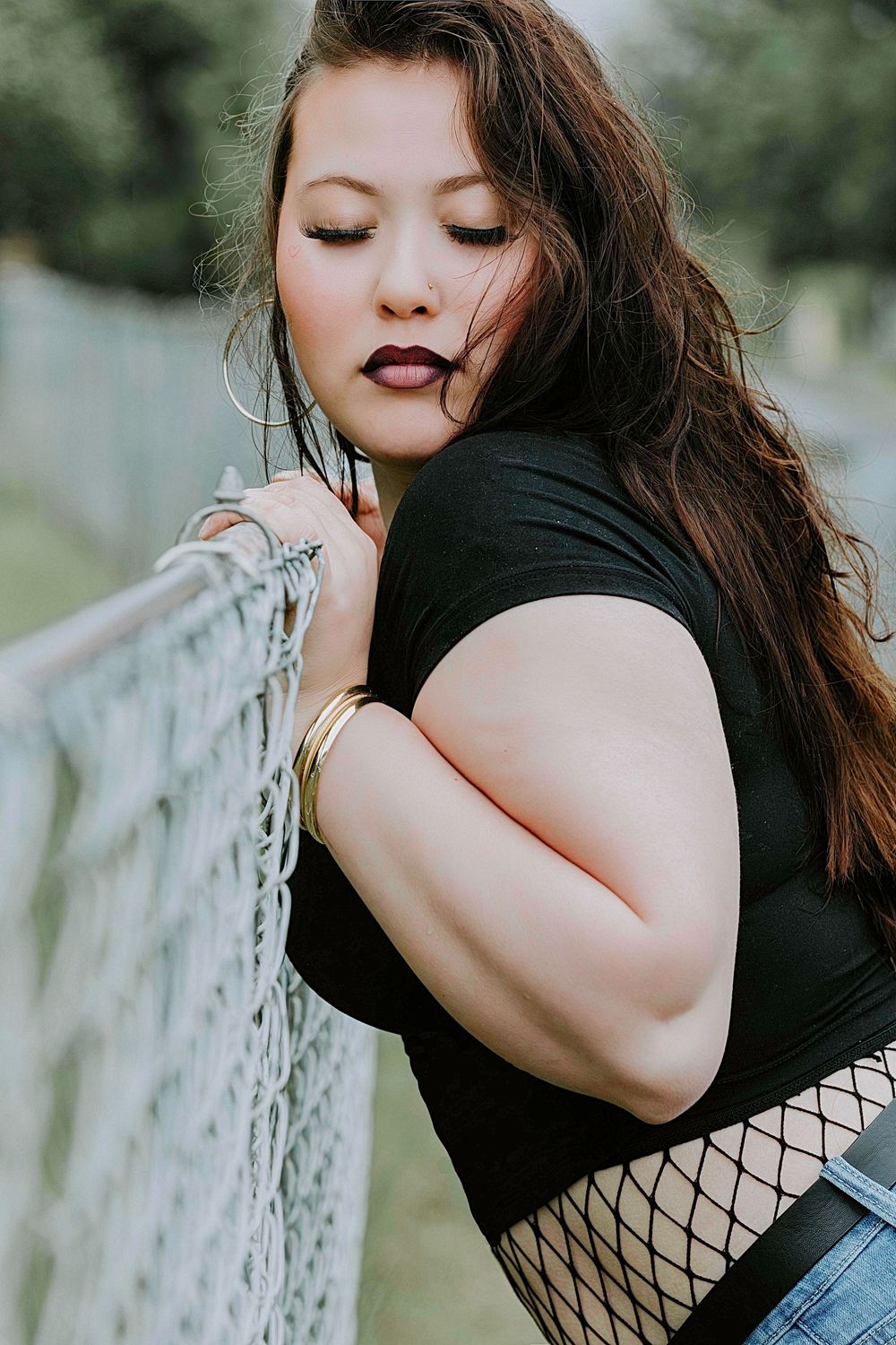 Artistic portrait of a person in a black top and fishnet stockings leaning against a railing in a natural setting.