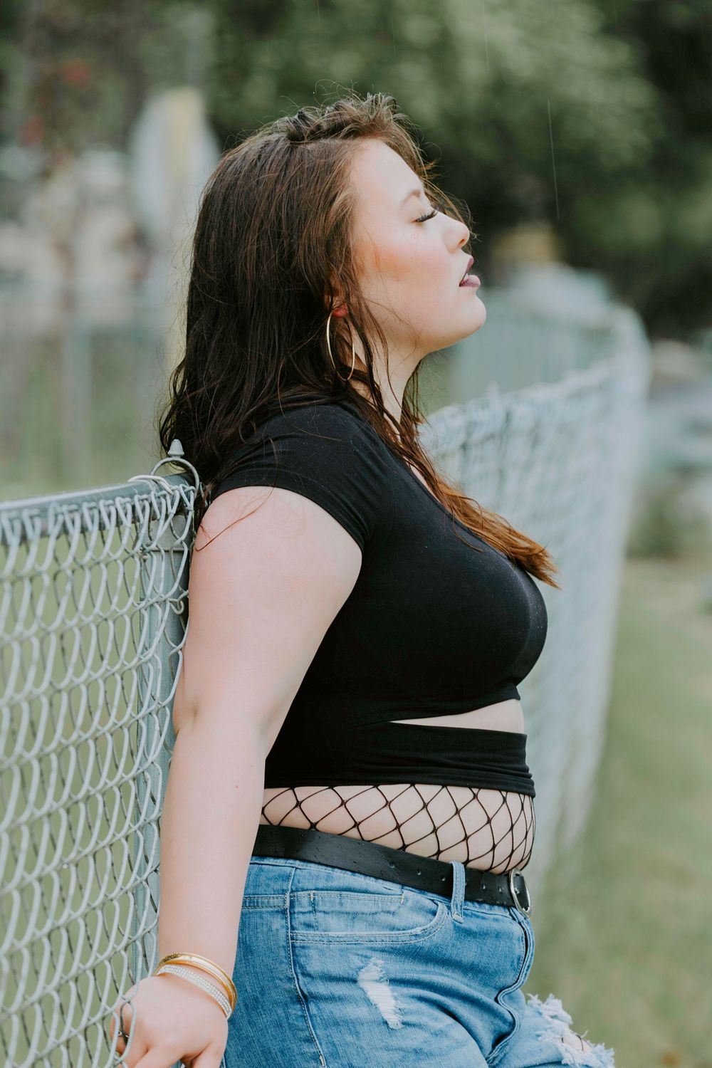 Fashion portrait of person in black crop top and ripped jeans leaning against chain link fence outdoors.