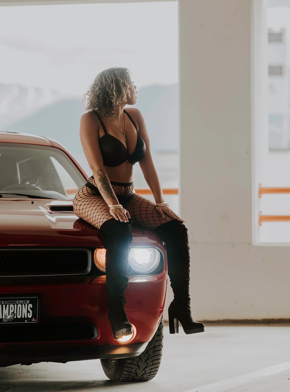 A red Dodge Challenger with illuminated headlights parked in a garage setting with someone sitting on the hood in dark attire.