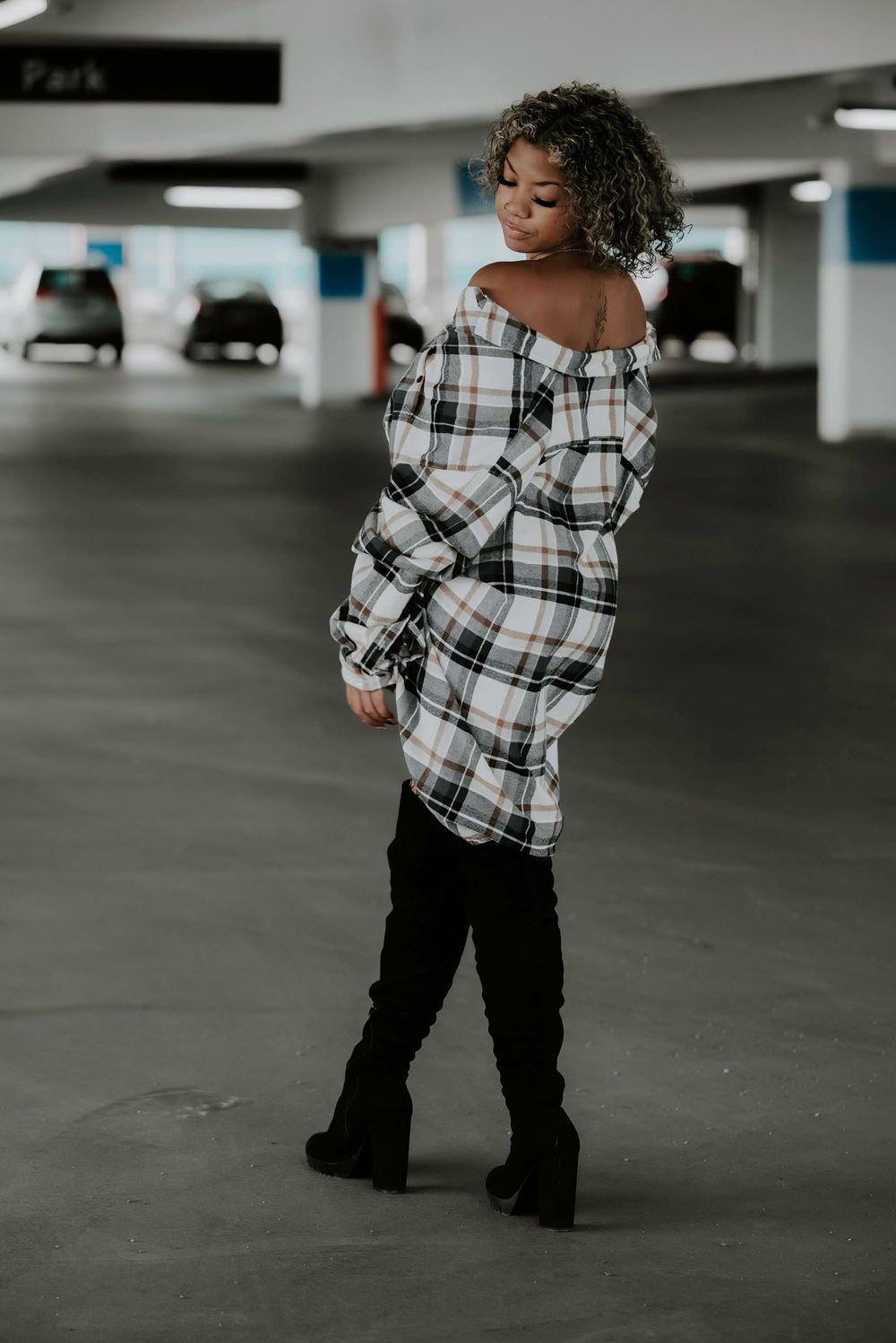 Someone in a stylish plaid off-shoulder dress and black thigh-high boots poses in a parking garage.