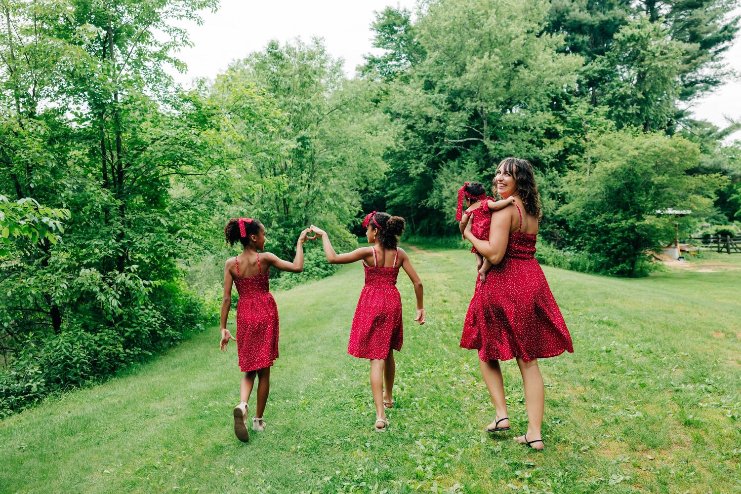 Three people in matching red dresses skip joyfully across a green lawn on a sunny day at Brooksvale Park in Hamden