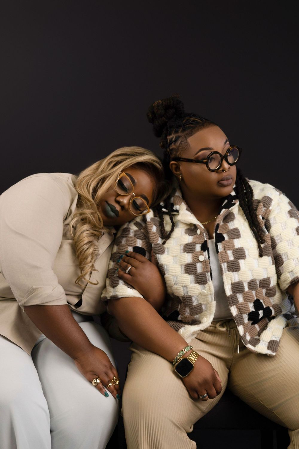 Two friends relaxing together on dark background wearing matching cream outfits with one wearing a patterned jacket.