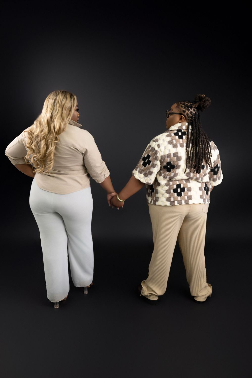 Two people wearing light colored casual clothing hold hands while standing with their backs to the camera against a dark background.