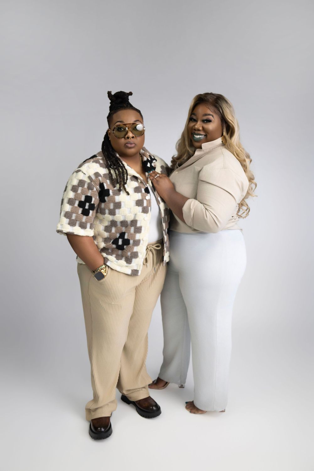 Two people in neutral toned outfits posing together against a gray backdrop for a fashion photoshoot.