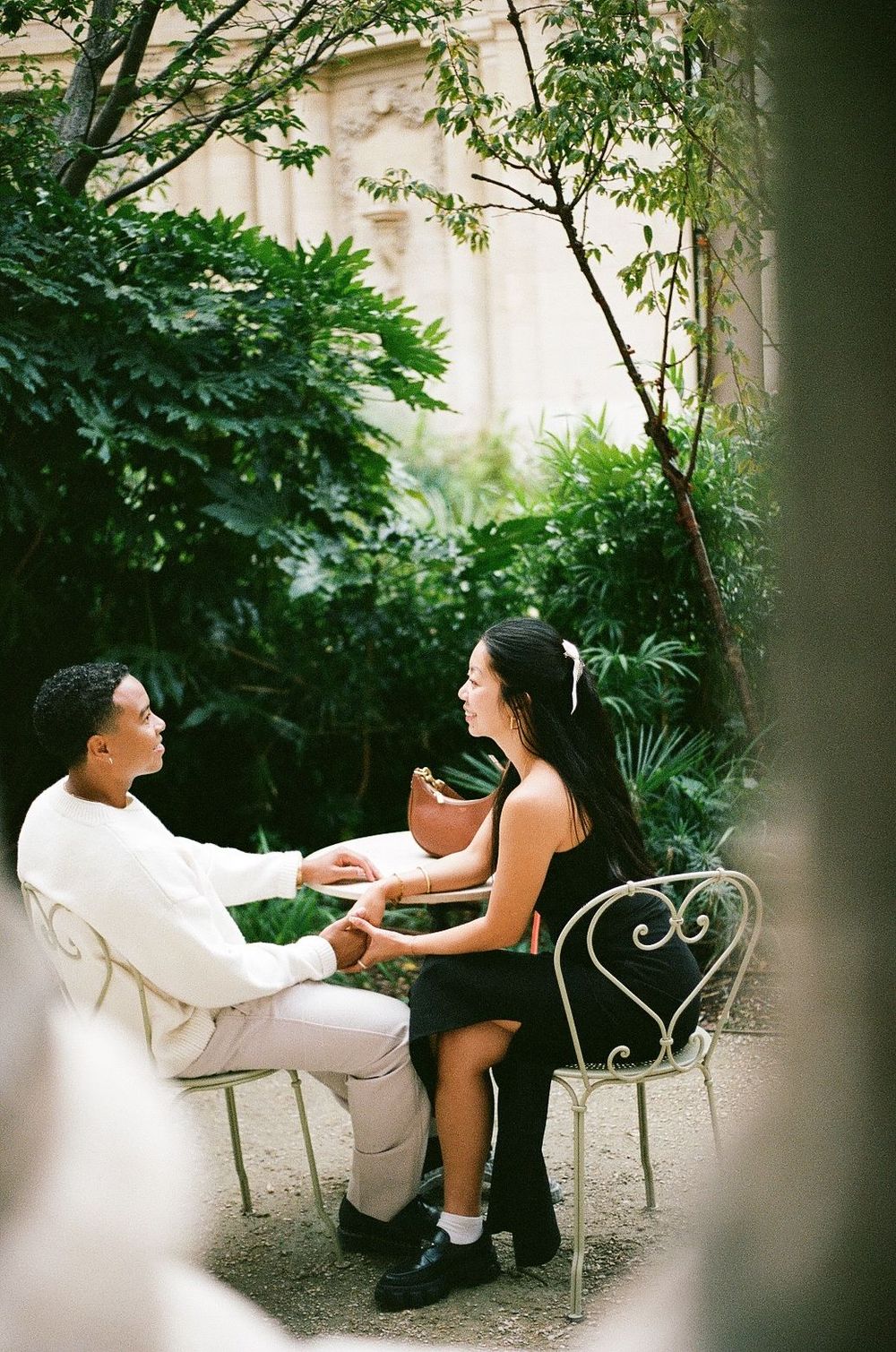 Two people sitting together on ornate white garden chairs in a lush green. Petit palais. Paris film photography.
