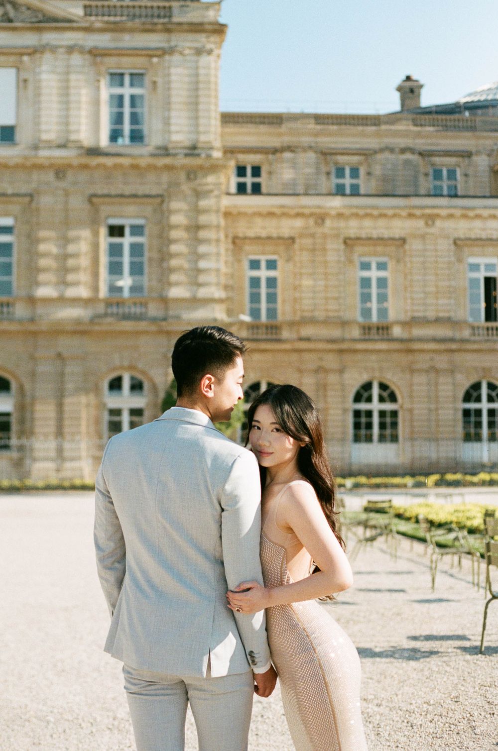 An elegant couple poses for a romantic pre-wedding photo in front of a grand French palace with beige stone architecture.
