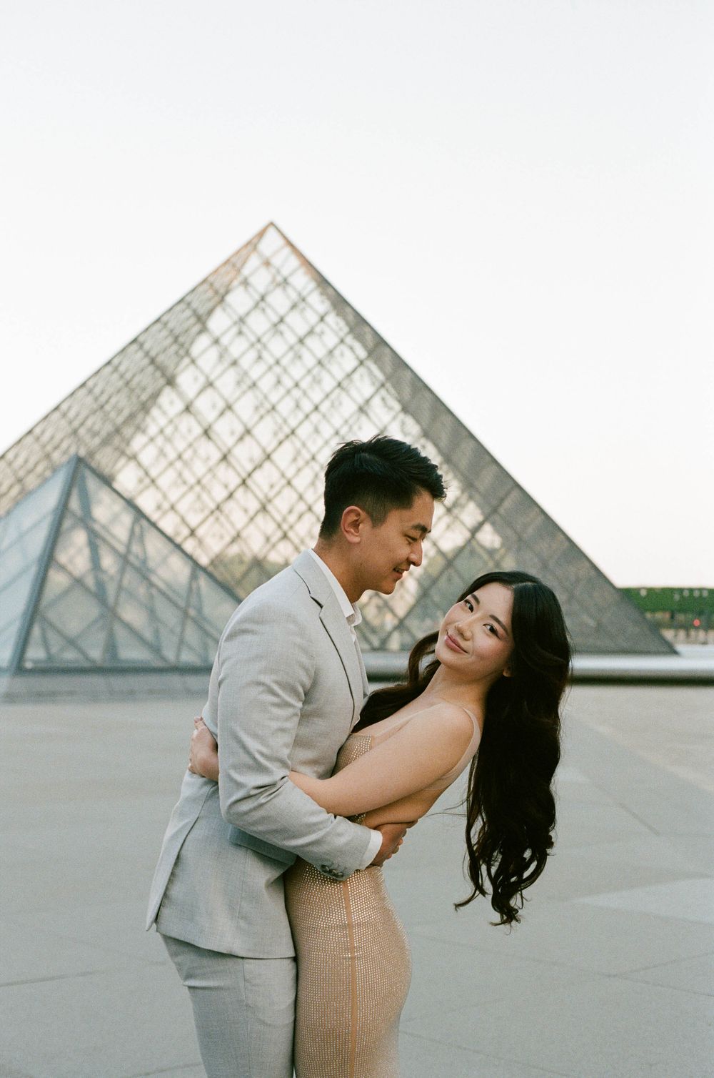 A romantic couple embraces in front of the iconic glass pyramid at the Louvre Museum in Paris.