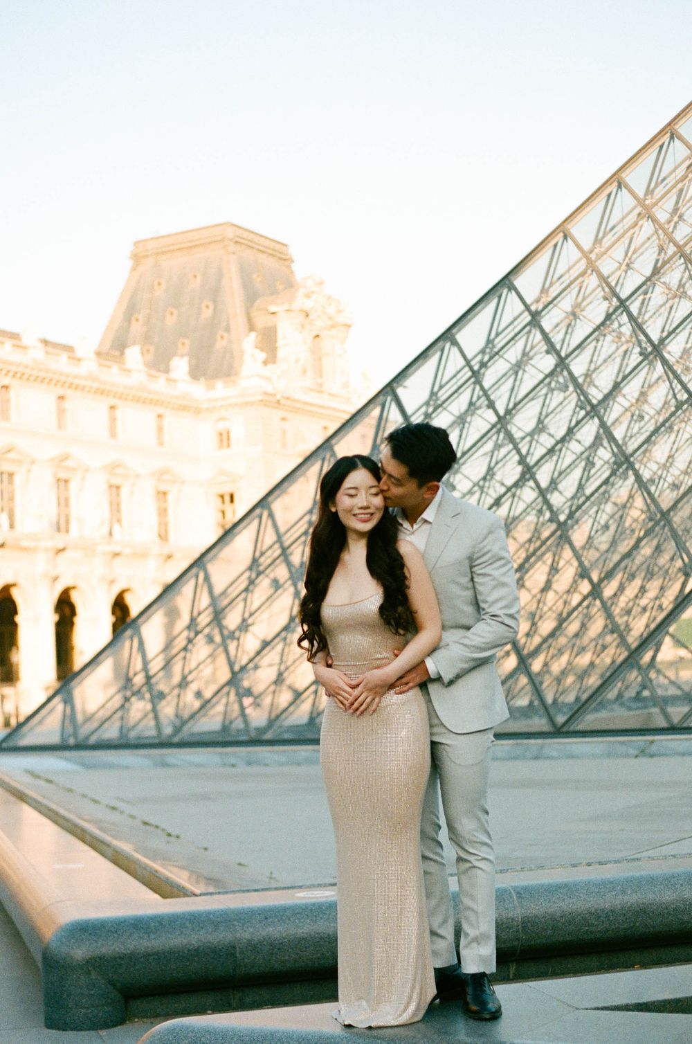 A couple sharing an intimate moment in front of the iconic glass pyramid at the Louvre Museum in Paris during sunset.
