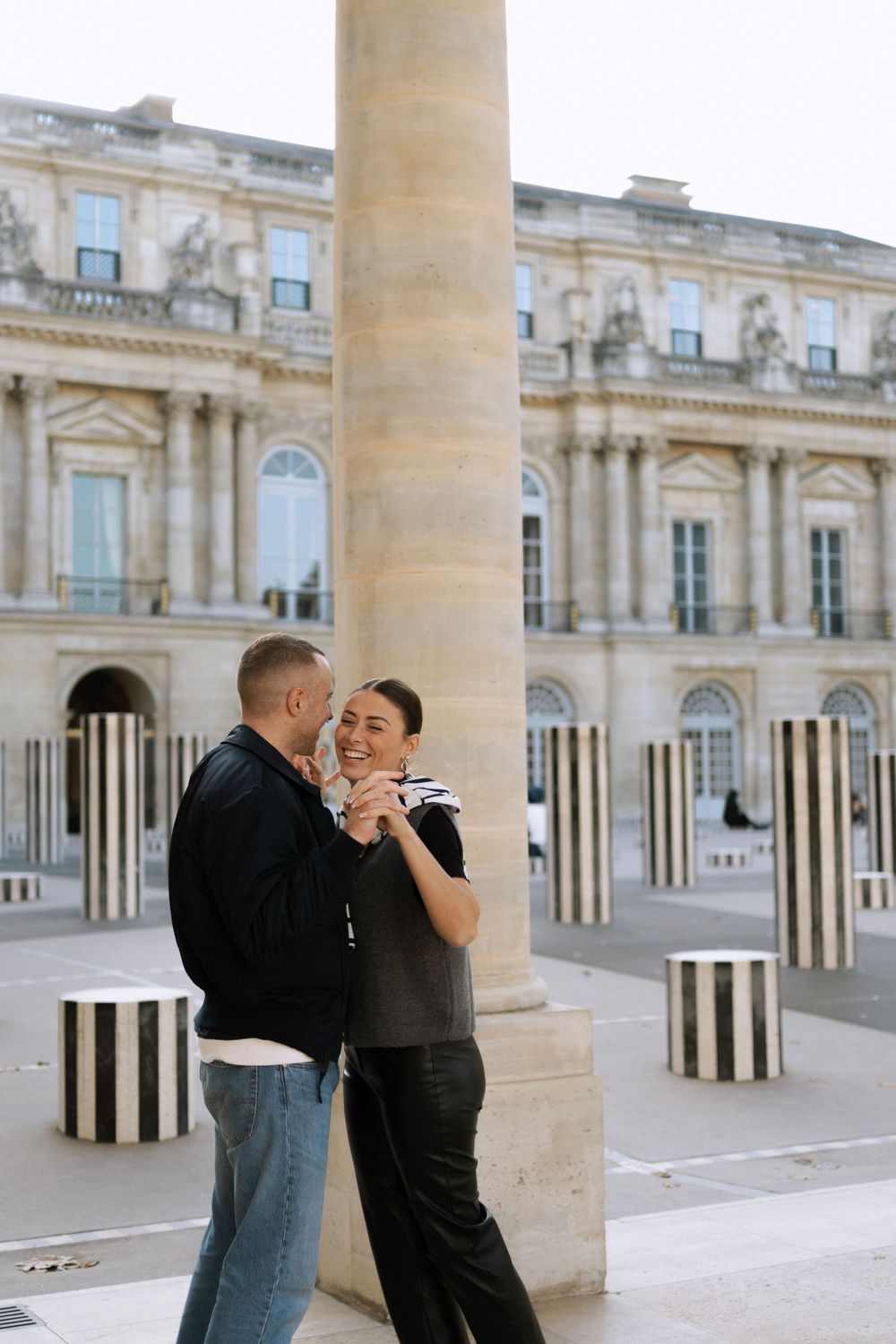 A romantic couple embraces near classical architecture columns with black and white striped bollards visible in Paris.