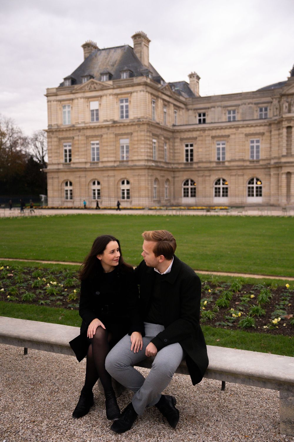 Two people sit and chat on a gravel path outside an elegant French chateau with manicured lawns.